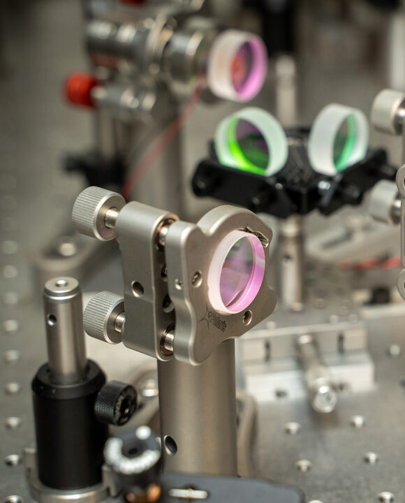 A close-up of a precision optical laboratory set up featuring multiple mounted lenses and mirrors with ping and green reflective coatings, arranged on a metallic optical bench for photonics experiments.